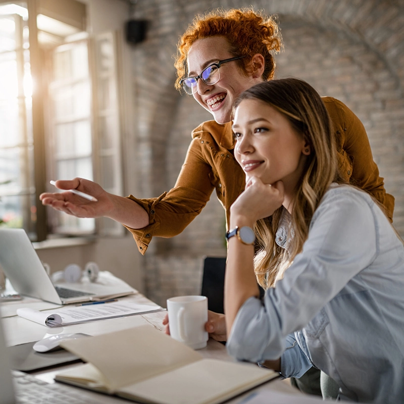 happy-female-entrepreneurs-reading-an-e-mail-on-computer-while-working-together-in-the-office-focus-is-on-redhead-woman- Happy female entrepreneurs reading an e-mail on computer while working together in the office. Focus is on redhead woman